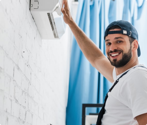 HVAC technician wearing a cap and overalls, smiling and installing a wall-mounted air conditioning unit in a bright room with blue curtains